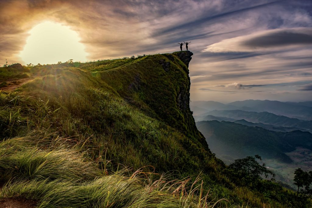 Zwei Personen im Sonnenuntergang stehen auf einem hervorstehenden Felsen und strecken jubelnd die Arme nach oben.