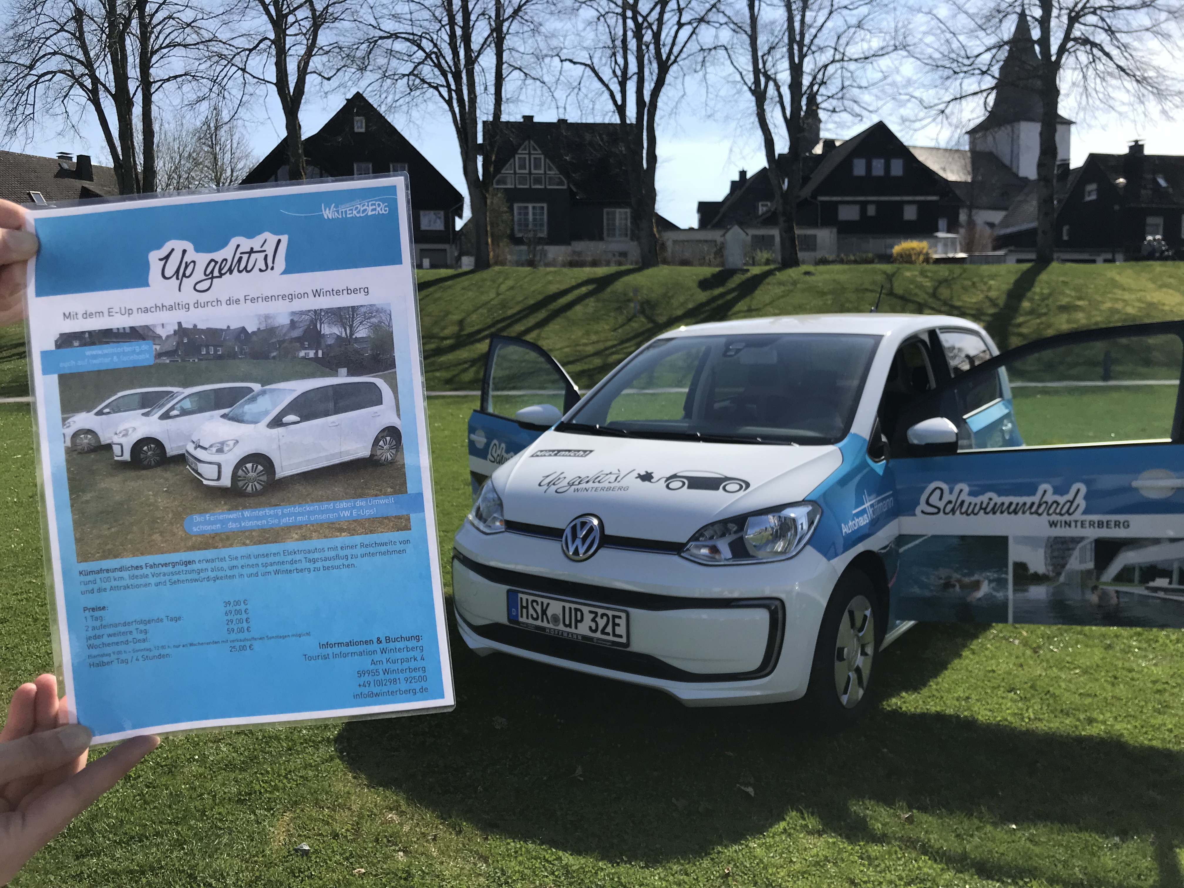 e-Auto (E-Up) das auf einer Wiese steht. eine Hand hält die Informationen und Preise für das e-Up in die Kamera.