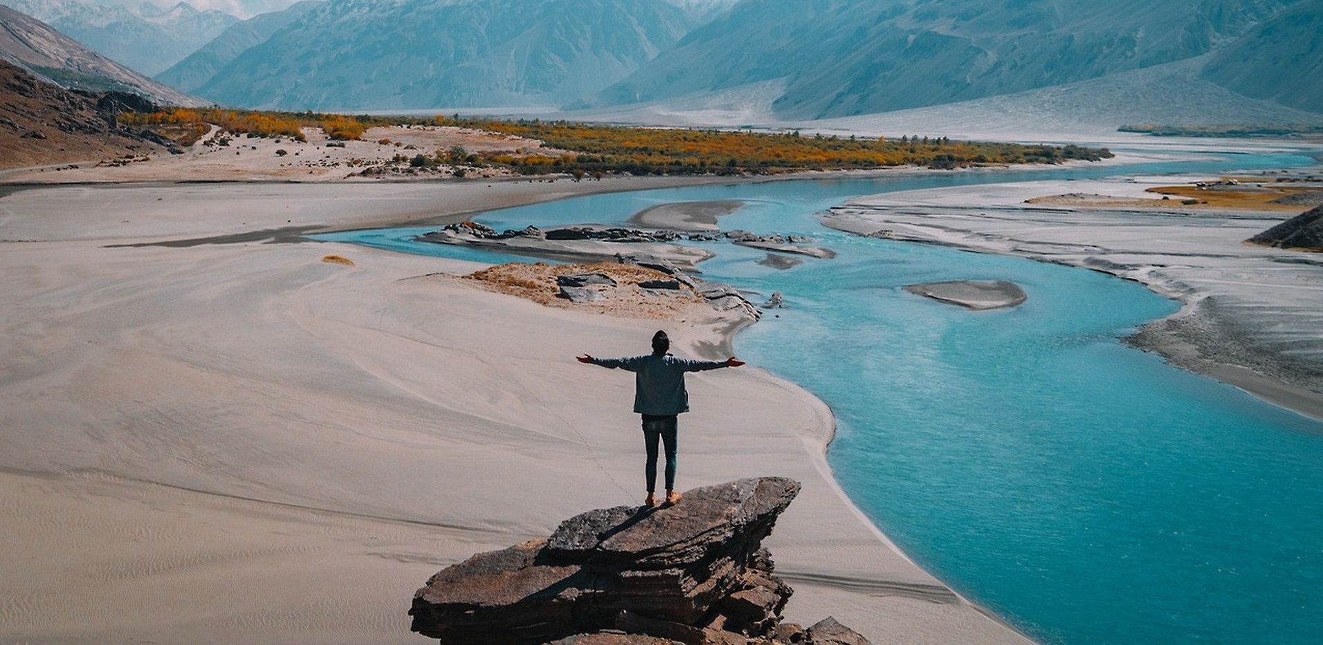 Person mit ausgestreckten Armen auf einem Stein stehend mit einer Berglandschaft und einem Fluss im Hintergrund