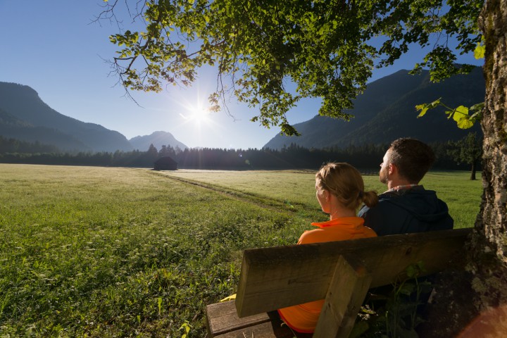 Ein Mann und eine Frau die bei sonnigem Wetter auf einer Bank sitzen und in die Berge schauen