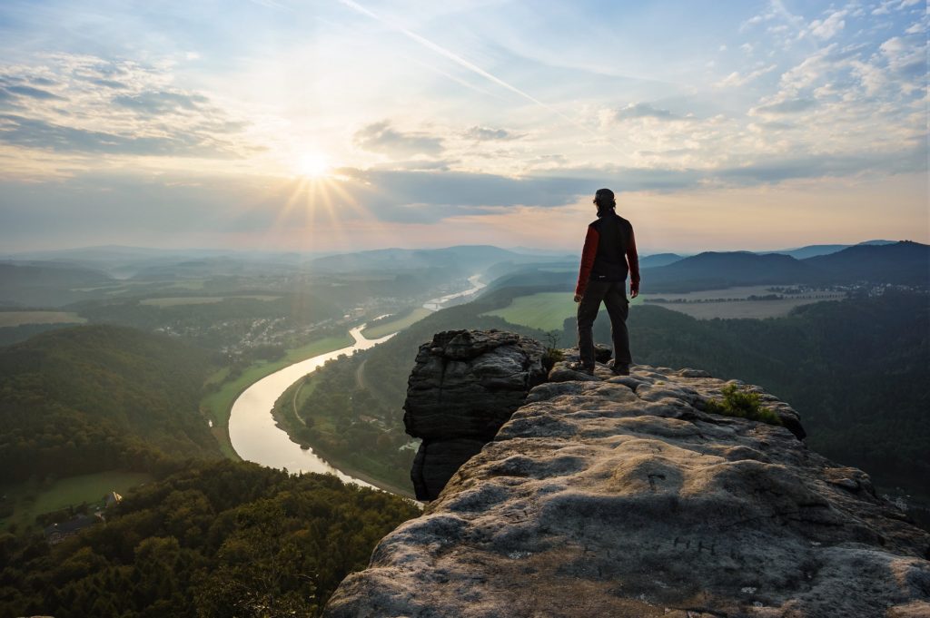 Die Silhouette eines Bergsteigers im Sonnenuntergang, der am Rand eines Felsens steht und in das Tal mit einem Fluss hinab blickt.