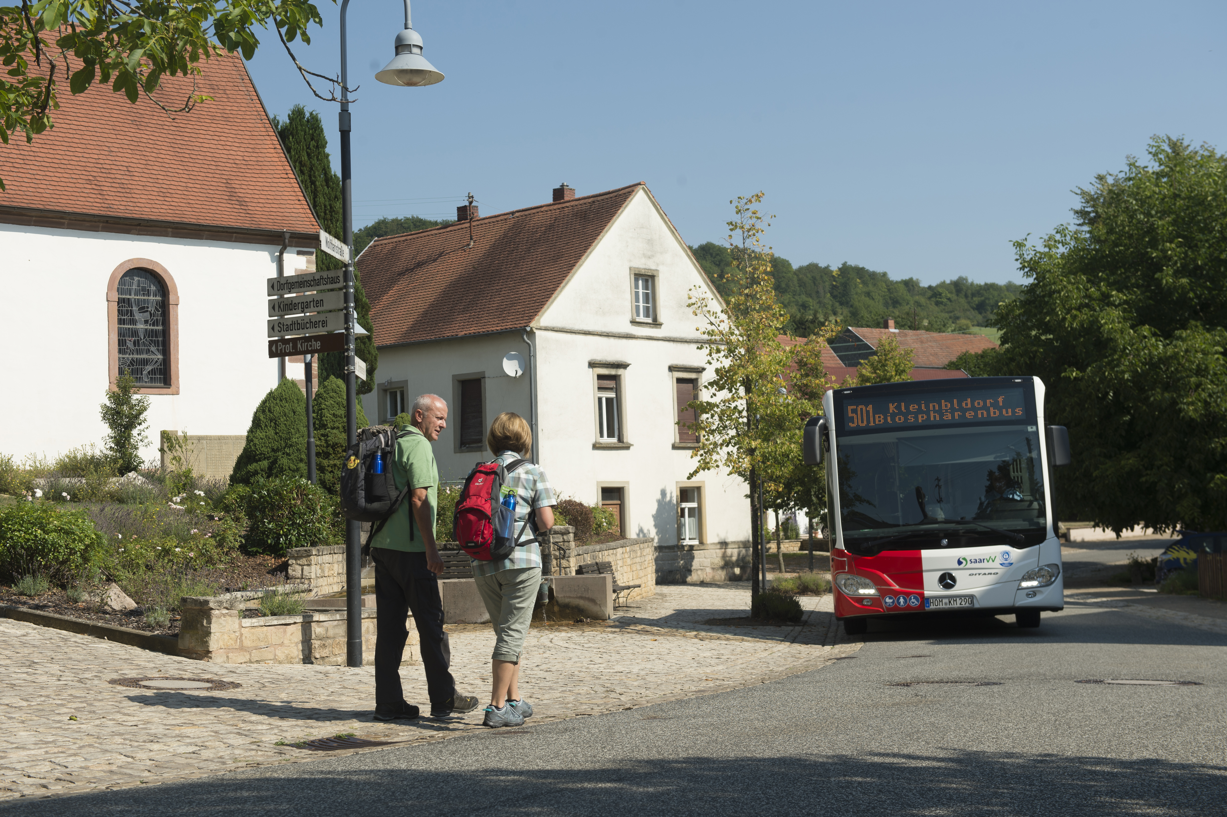 ein Mann und eine Frau mit Rucksäcken auf den Schultern auf dem Weg zu einer Bushaltestelle an der ein Bus steht