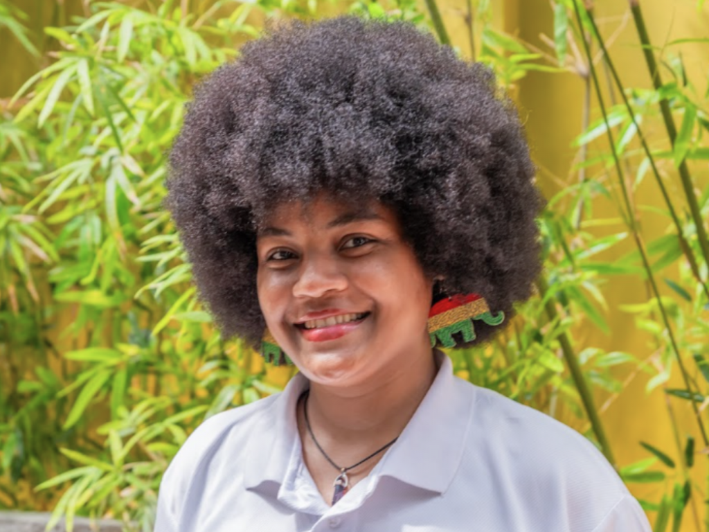 Empleada de hotel con una camisa tipo polo blanca, de pie frente a unas plantas y mirando directamente a la cámara.